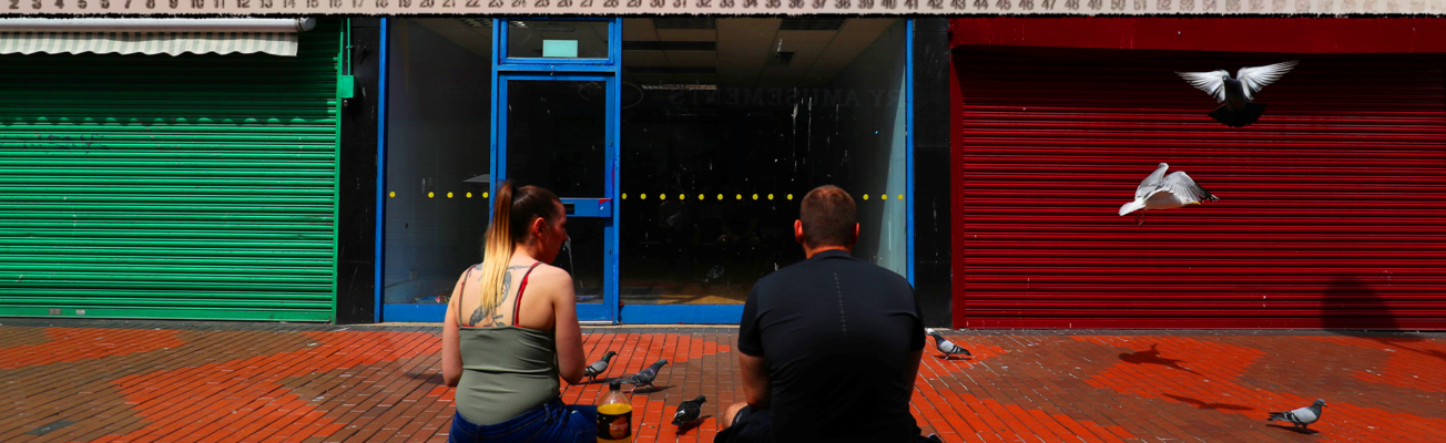 Two people sat facing shuttered high street shops.