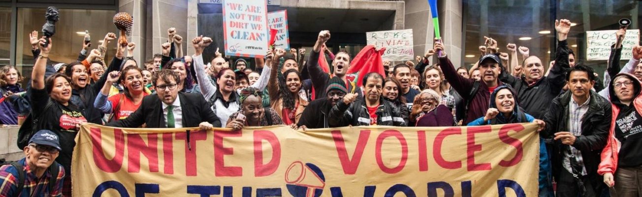 A group of workers stand with raised fists behind a banner that reads 'United Voices of the World'