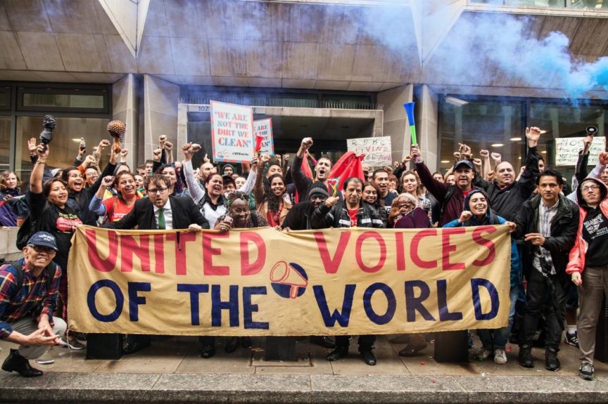 A group of workers stand with raised fists behind a banner that reads 'United Voices of the World'
