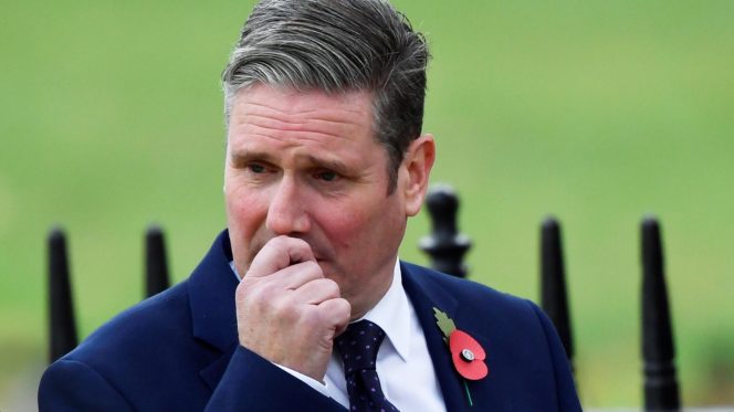 Keir Starmer wearing a suit and red poppy and looking worried as he holds his hand to his mouth