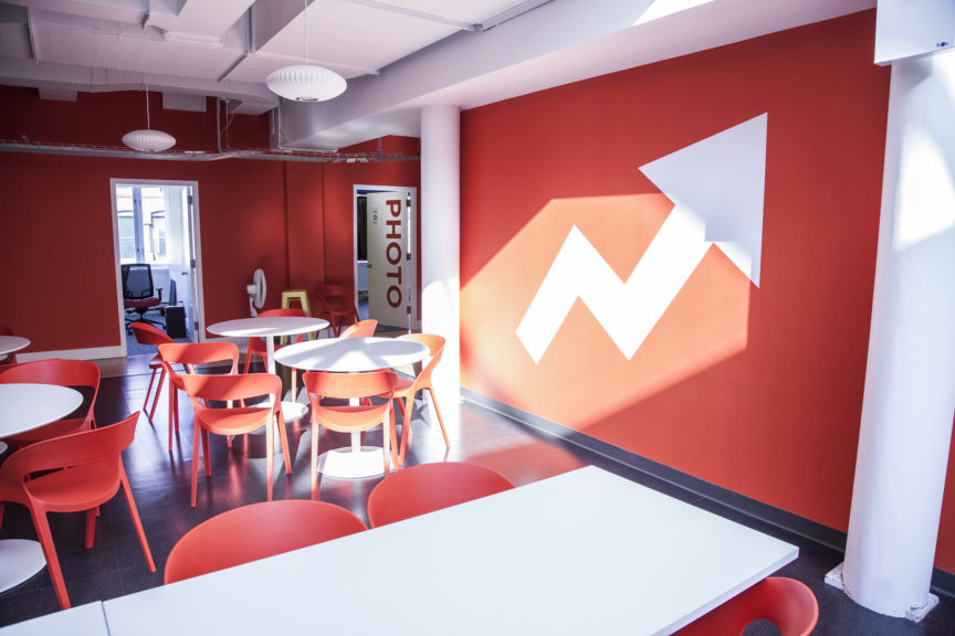 A picture of the offices of BuzzFeed, with white tables and red chairs, and a red wall with the company's distinctive white arrow