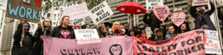 image of a crowd of protestors in london with pink and red signs reading "sex work is work", "outlaw poverty not prostitutes", "rights not raids" and "soho says: safety for sex workers"