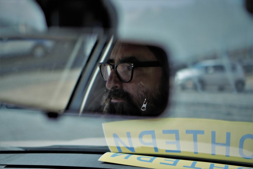 A man is seen in the mirror of a car. The sign 'Volunteer' is wedged on the dashboard.