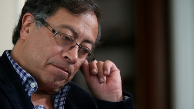 Image shows a close up portrait of new Colombian president Gustavo Petro, biting his lip, his left hand raised and twiddling his ear. He is wear a pair of glasses, a checked blue shirt and a dark blue jacket.