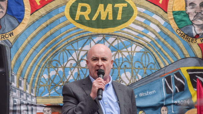 Images shows RMT union General Secretary Mick Lynch speaks during the rally outside King's Cross Station. He is wearing a grey suit with a blue shirt, standing in front of a vibrant embroidered banner with RMT sewed on it in yellow thread.