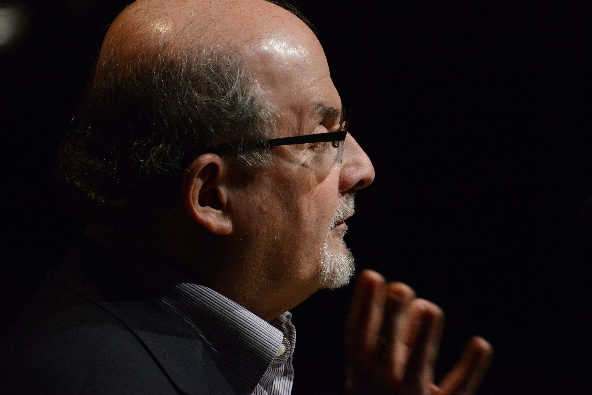 A bald man with black-grey sideburns and glasses seen close up in profile, with his hand raised by his face