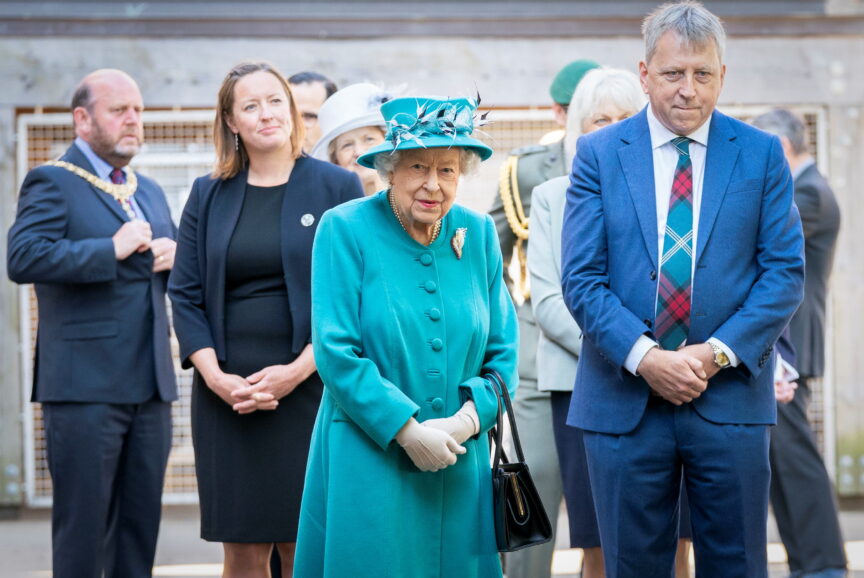 A man in a blue suit stands besides the late Queen, who is wearing a turqoise outfit