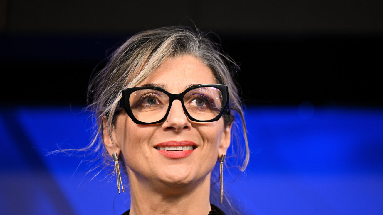 A woman with grey hair and glasses smiles at an audience