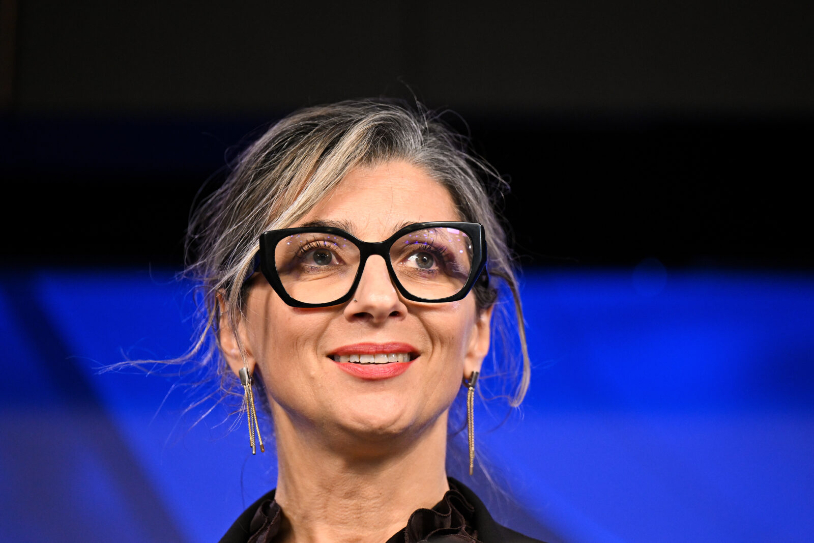 A woman with grey hair and glasses smiles at an audience