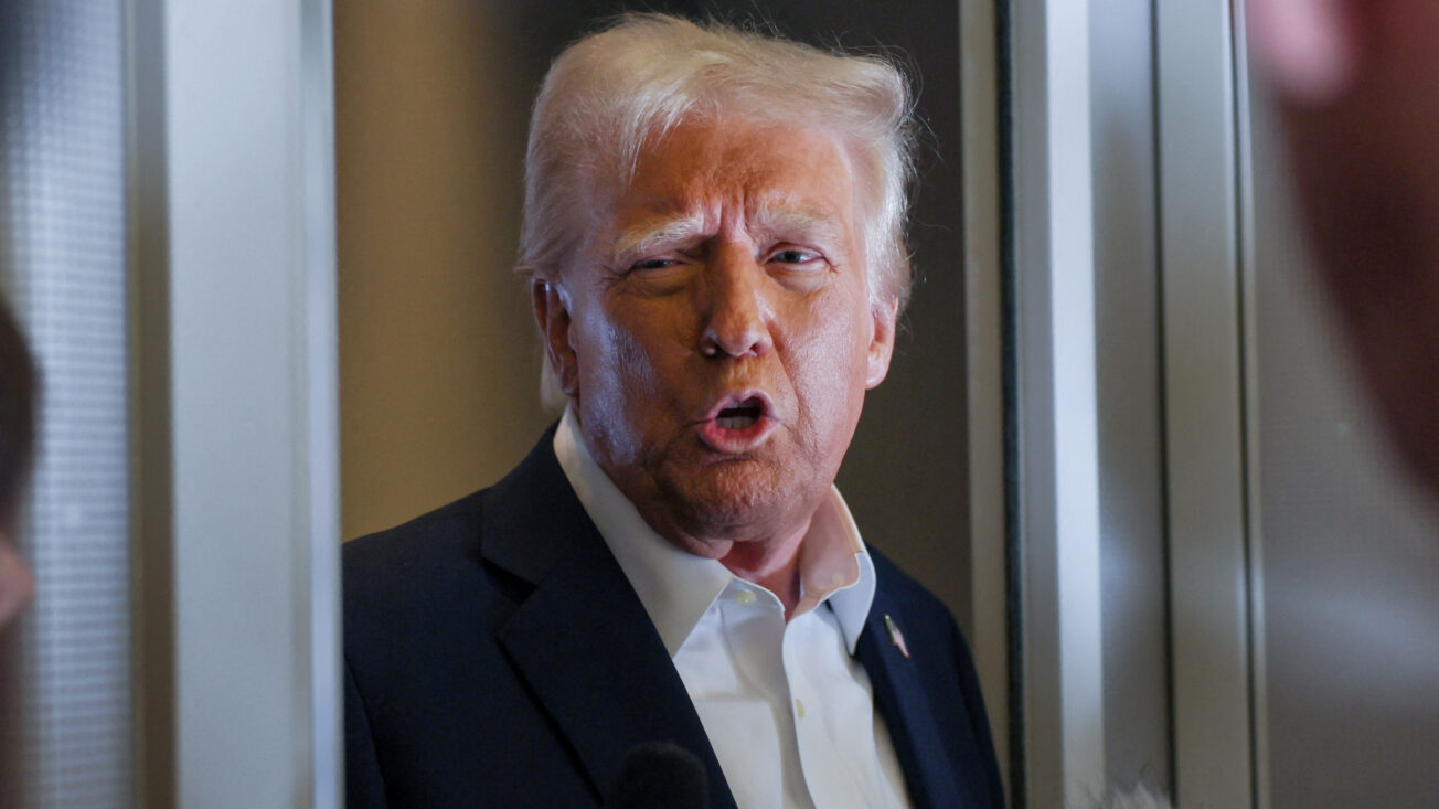 A man in a suit with blonde hair speaks with microphones underneath him held by reporters