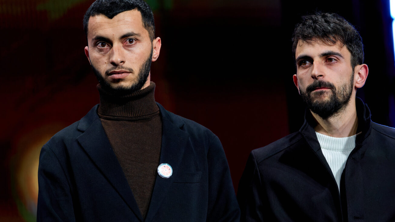 Two men in suits stand next to one another looking stern