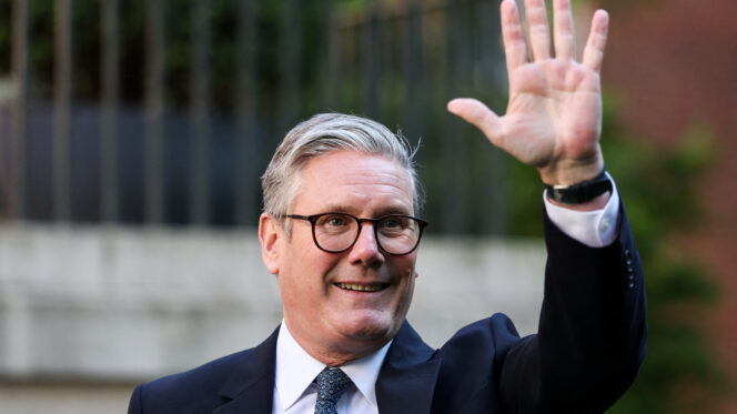 A man with silver hair and glasses, wearing a suit, waves a hand in the air