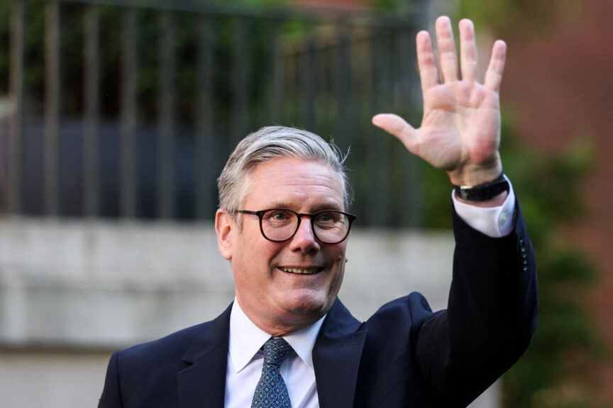 A man with silver hair and glasses, wearing a suit, waves a hand in the air
