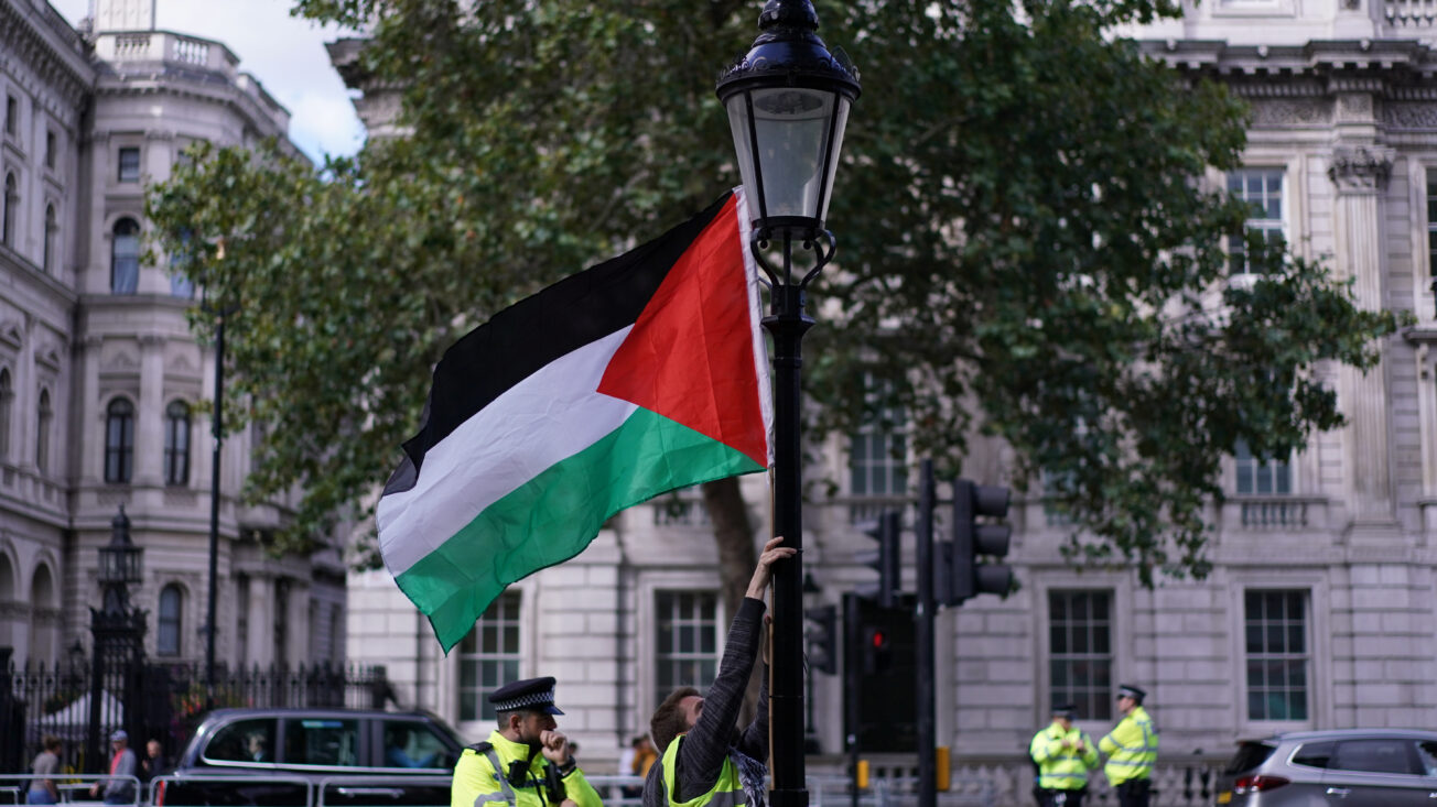 A man in a yellow tabard puts up a Palestine flag near a street lamp with large white buildings in the background