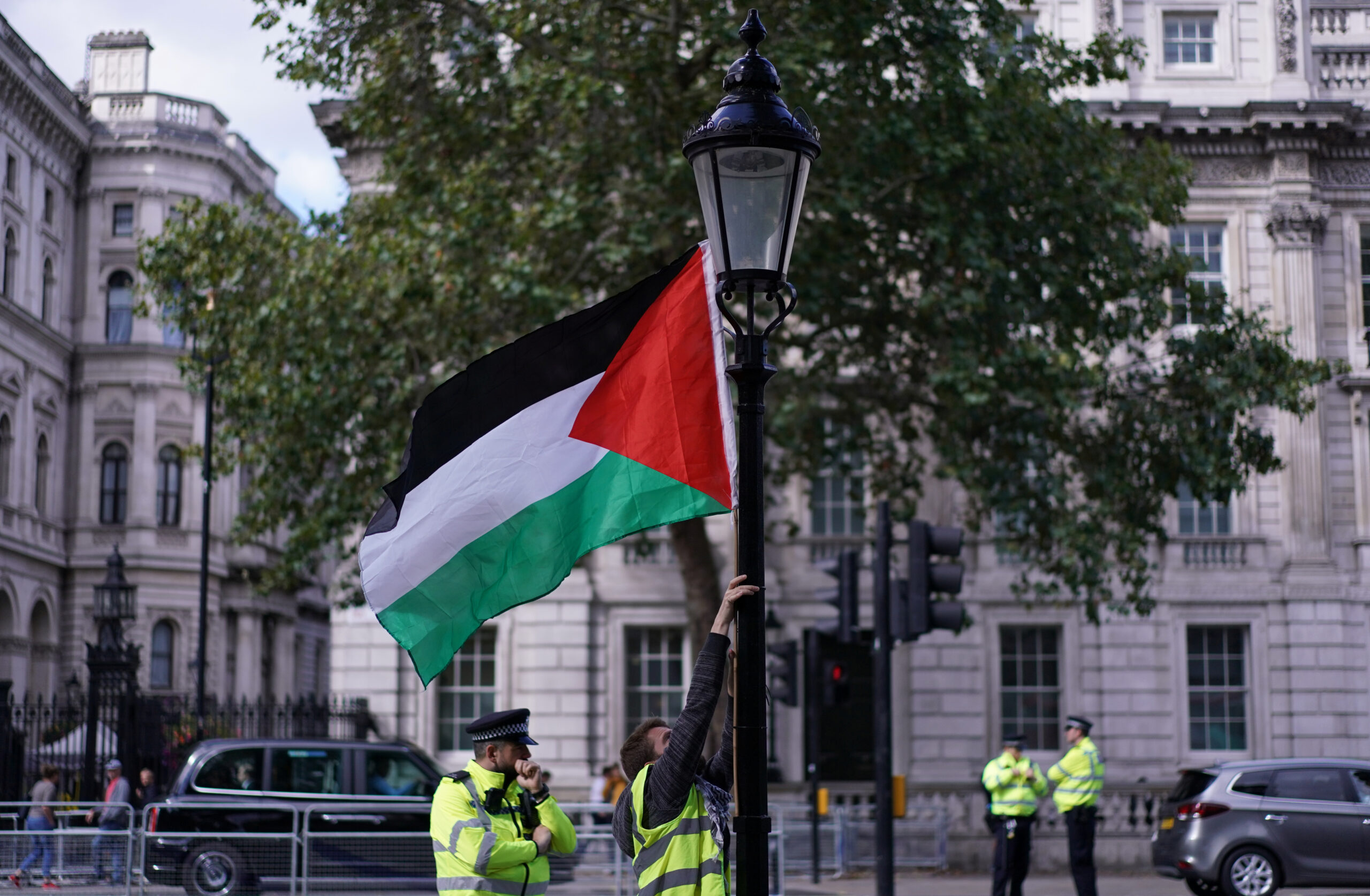 A man in a yellow tabard puts up a Palestine flag near a street lamp with large white buildings in the background