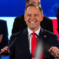 A man with red face, short blondish grey hair, a black suit and red tie holds two microphones as people clap behind him