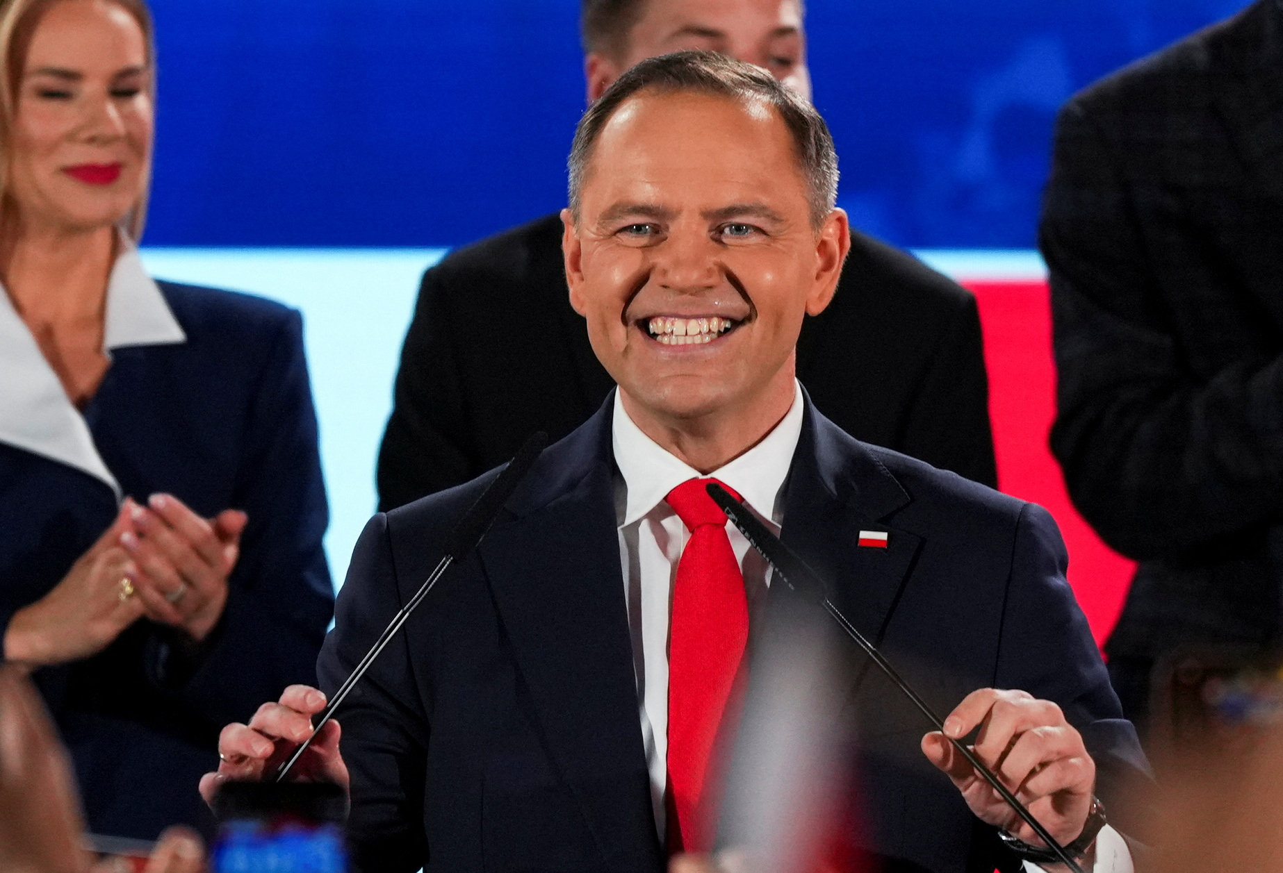 A man with red face, short blondish grey hair, a black suit and red tie holds two microphones as people clap behind him