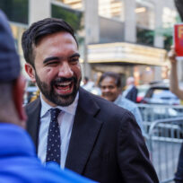 A man smiles in foreground with a sign saying 'freeze the rent' in the background