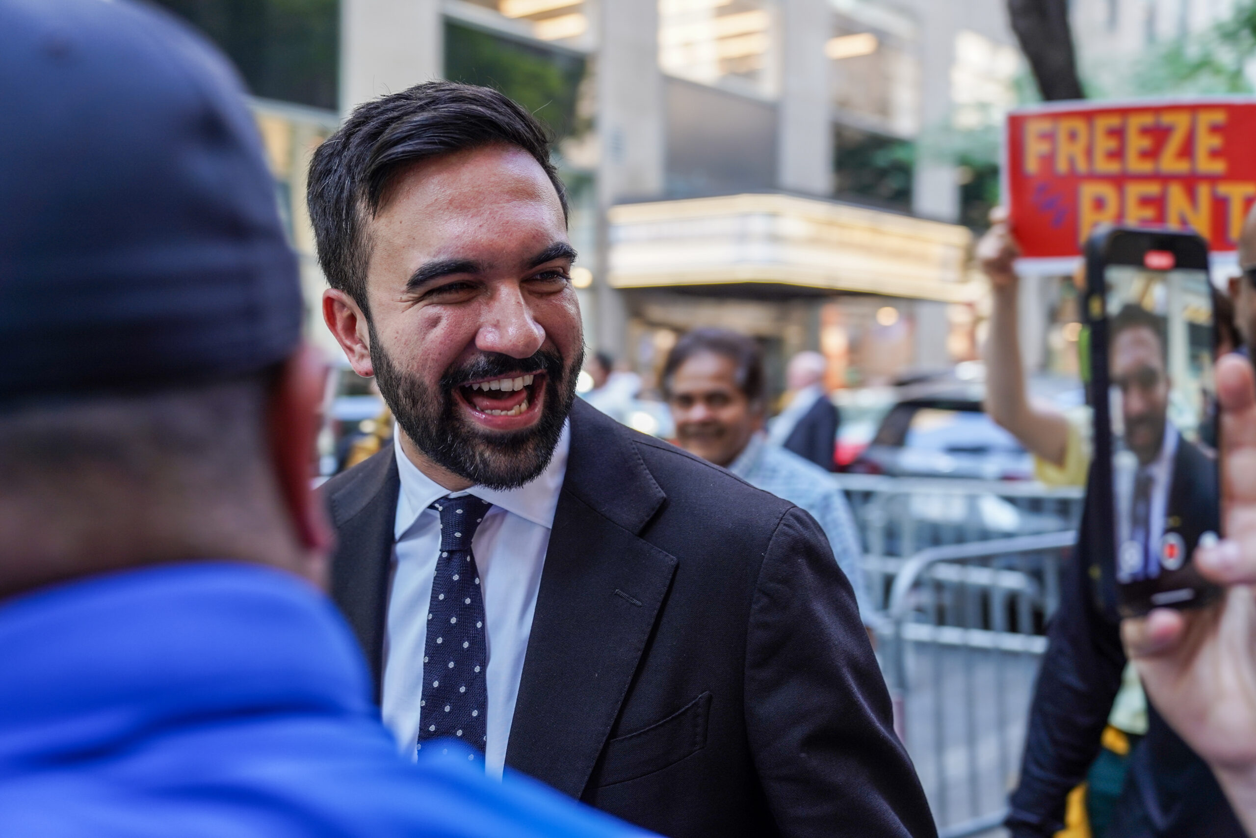 A man smiles in foreground with a sign saying 'freeze the rent' in the background