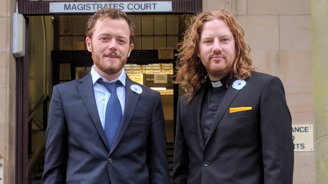 Two men in suits stand outside a court building. One wears a dog collar.