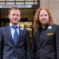 Two men in suits stand outside a court building. One wears a dog collar.