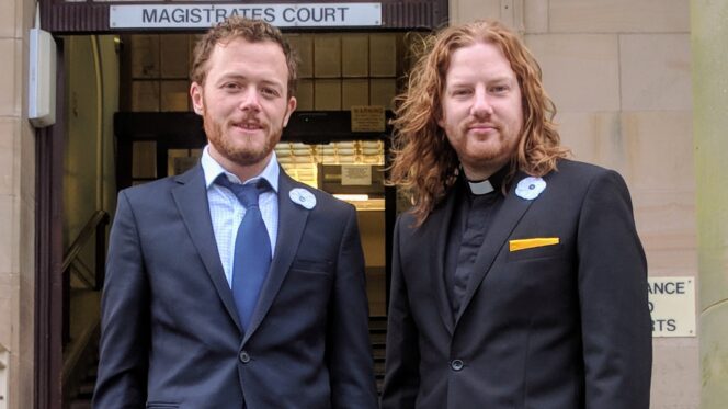 Two men in suits stand outside a court building. One wears a dog collar.