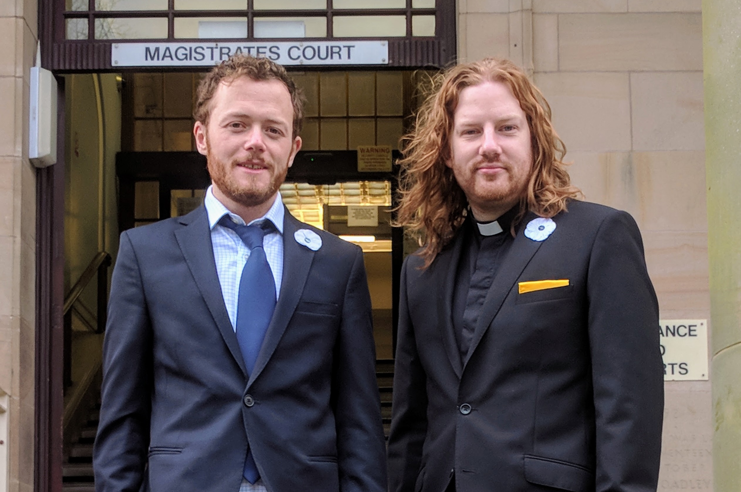 Two men in suits stand outside a court building. One wears a dog collar.