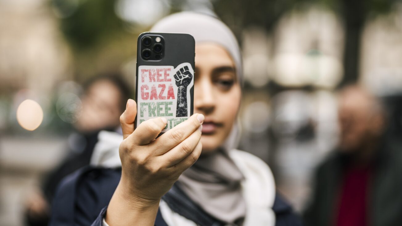 A woman in a grey hijab holds up her phone to take a photograph. The phone has a sticker on the back that says "Free Gaza".