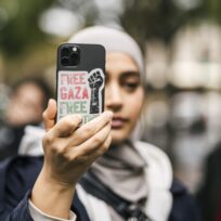 A woman in a grey hijab holds up her phone to take a photograph. The phone has a sticker on the back that says "Free Gaza".