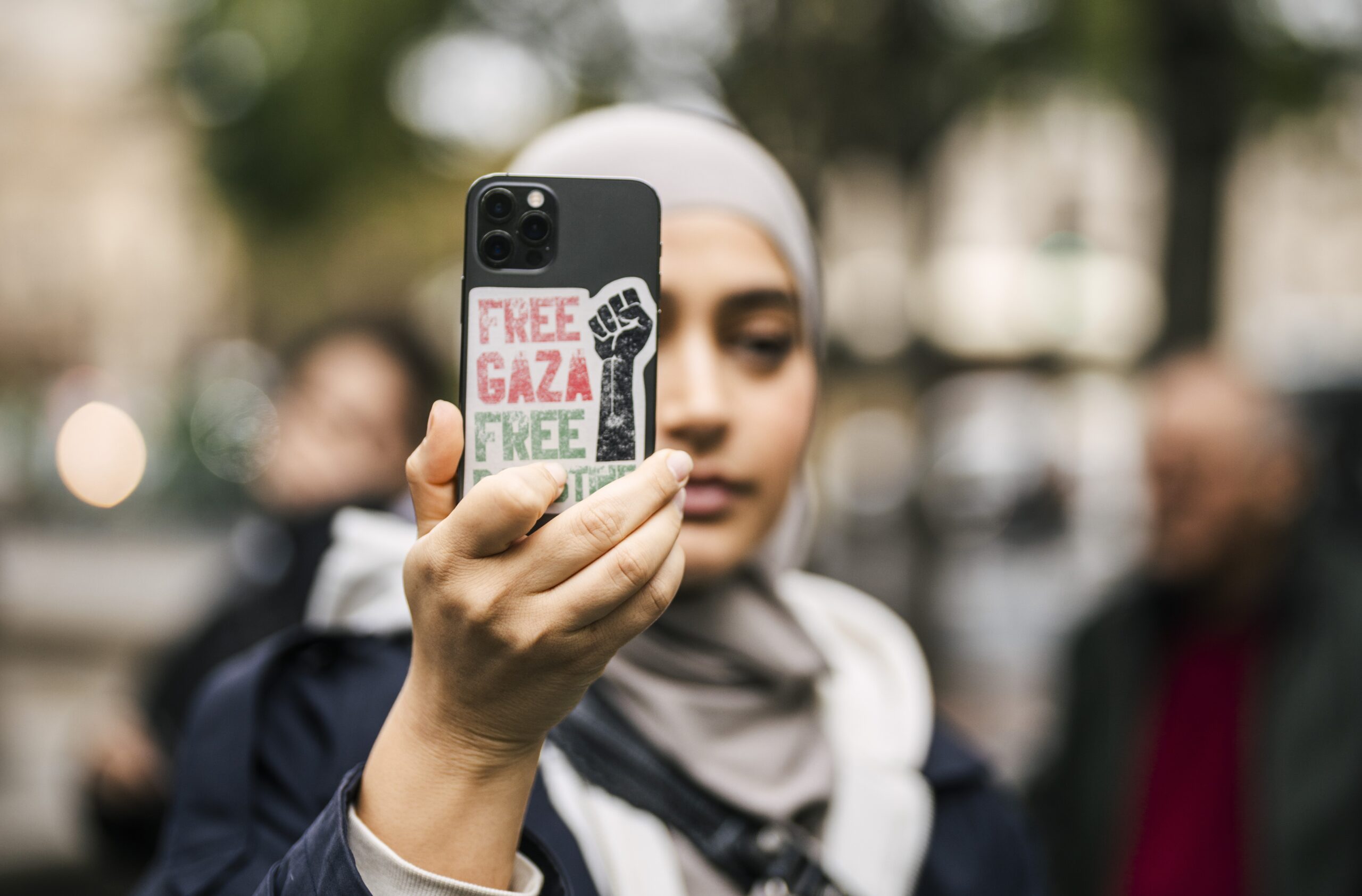 A woman in a grey hijab holds up her phone to take a photograph. The phone has a sticker on the back that says "Free Gaza".