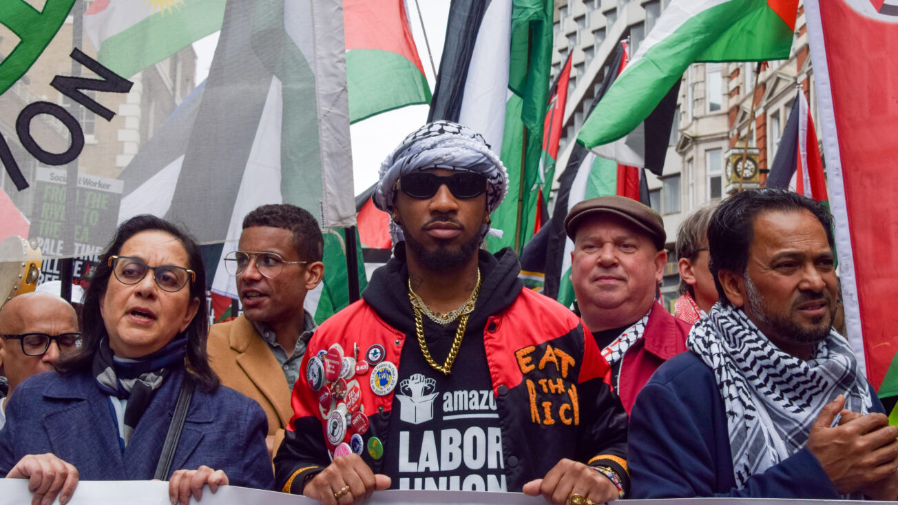 A Black man wearing a keffiyeh as a headscarf holds a banner alongside Palestinian flags