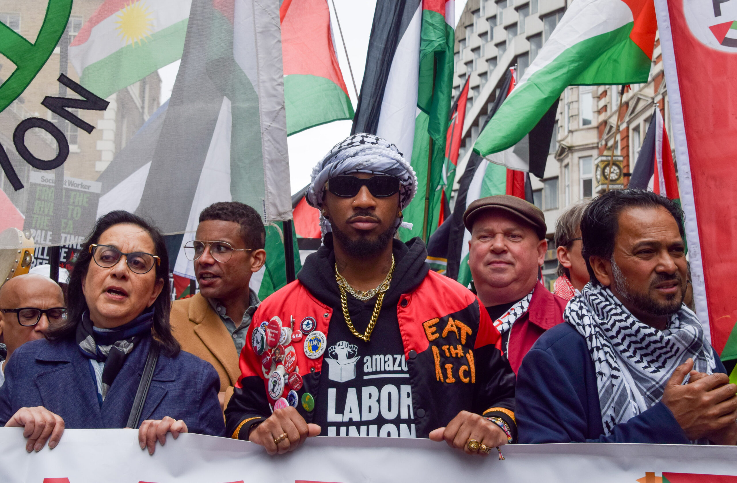 A Black man wearing a keffiyeh as a headscarf holds a banner alongside Palestinian flags