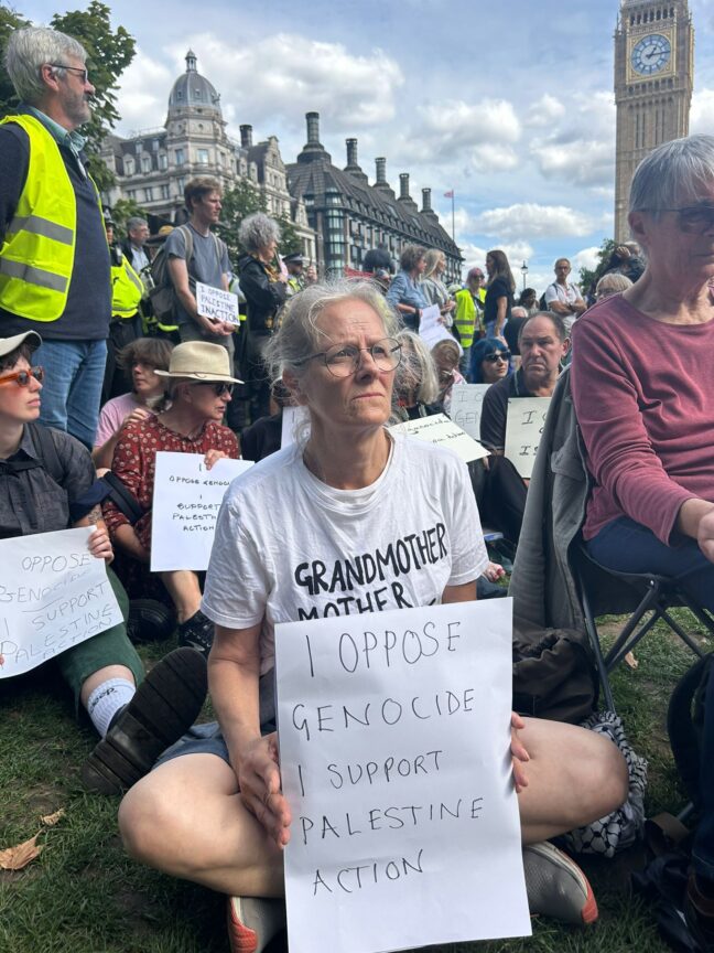 Saturday 6 August: An older woman in Parliament Square holds a paper sign that reads: “I oppose genocide. I support Palestine Action.”