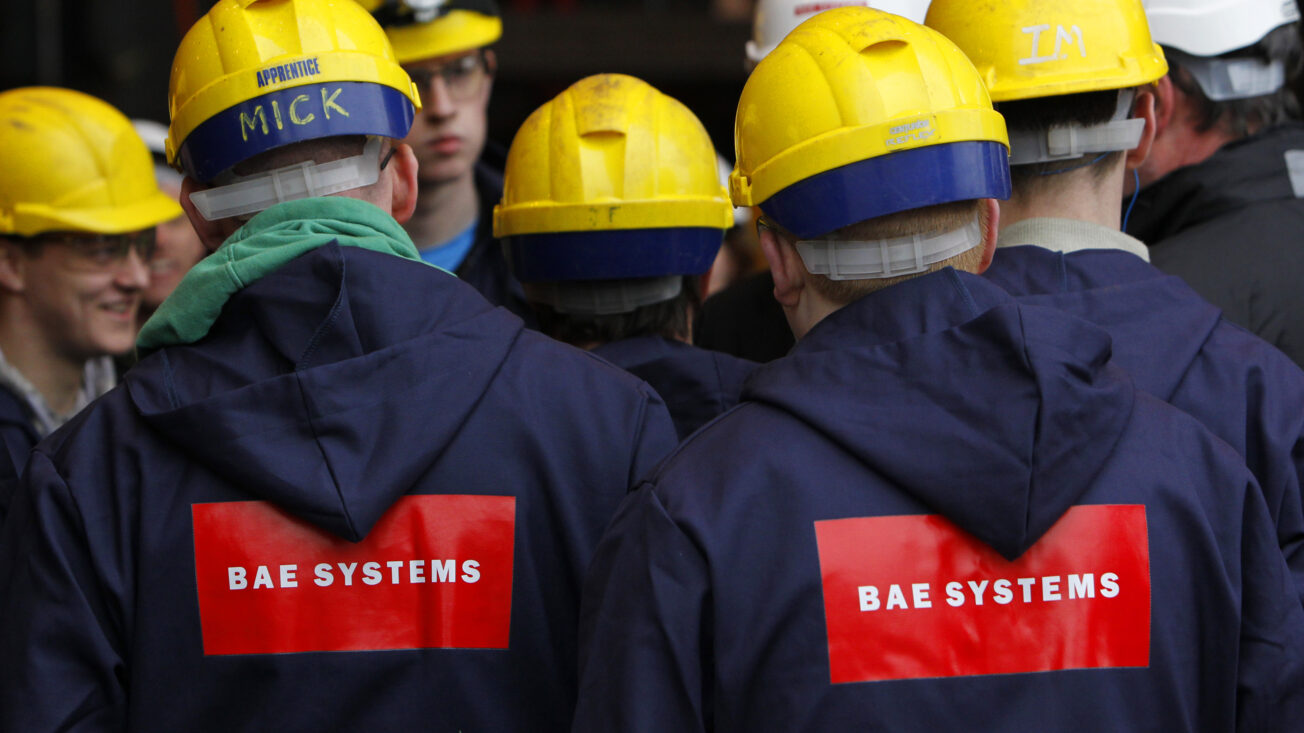 A group of people in yellow hard hats with their backs to the camera wear navy overalls with the logo 'BAE Systems' on them
