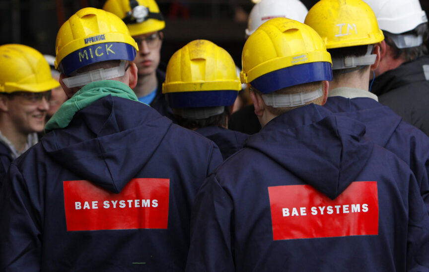 A group of people in yellow hard hats with their backs to the camera wear navy overalls with the logo 'BAE Systems' on them