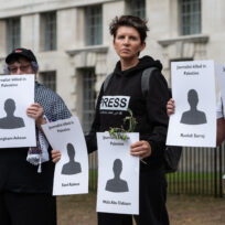 A person with a black hoodie that says 'press' on it holds a piece of card that has a cut-out figure with the words 'journalist killed in palestine', and the name of a killed Gazan journalist.