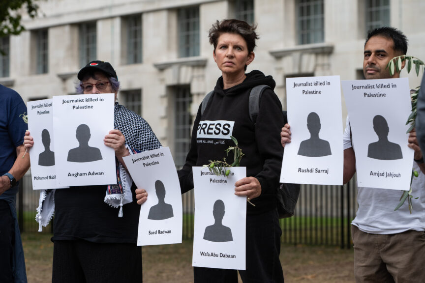 A person with a black hoodie that says 'press' on it holds a piece of card that has a cut-out figure with the words 'journalist killed in palestine', and the name of a killed Gazan journalist.