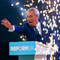 A man with grey hair and a navy suit outstretches his hand at a podium that says 'Reform UK' as fireworks go off around him