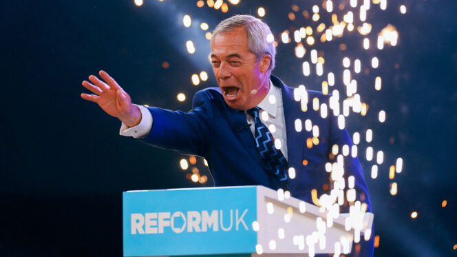 A man with grey hair and a navy suit outstretches his hand at a podium that says 'Reform UK' as fireworks go off around him
