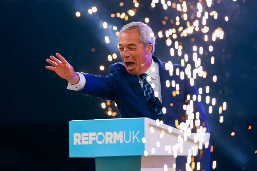 A man with grey hair and a navy suit outstretches his hand at a podium that says 'Reform UK' as fireworks go off around him