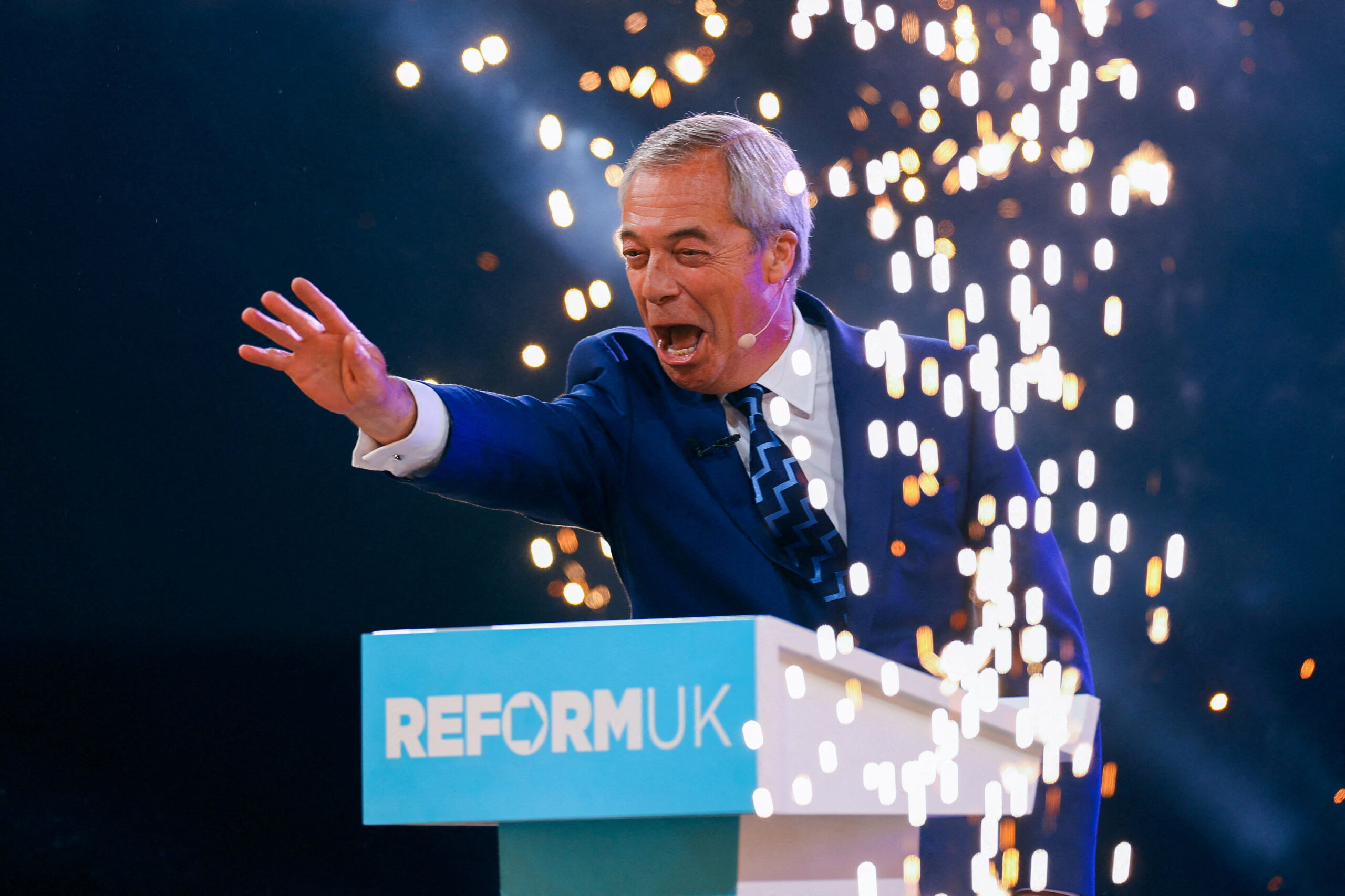 A man with grey hair and a navy suit outstretches his hand at a podium that says 'Reform UK' as fireworks go off around him