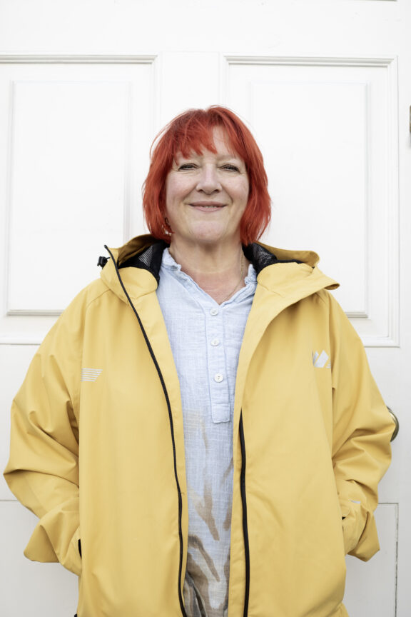 A woman with red hair and a yellow raincoat stands in front of a door