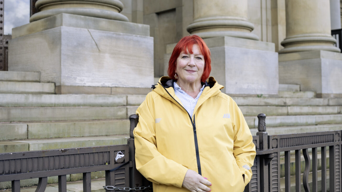 A woman with red hair and a yellow raincoat stands in front of railings in front of a neoclassical building