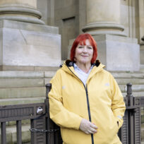 A woman with red hair and a yellow raincoat stands in front of railings in front of a neoclassical building