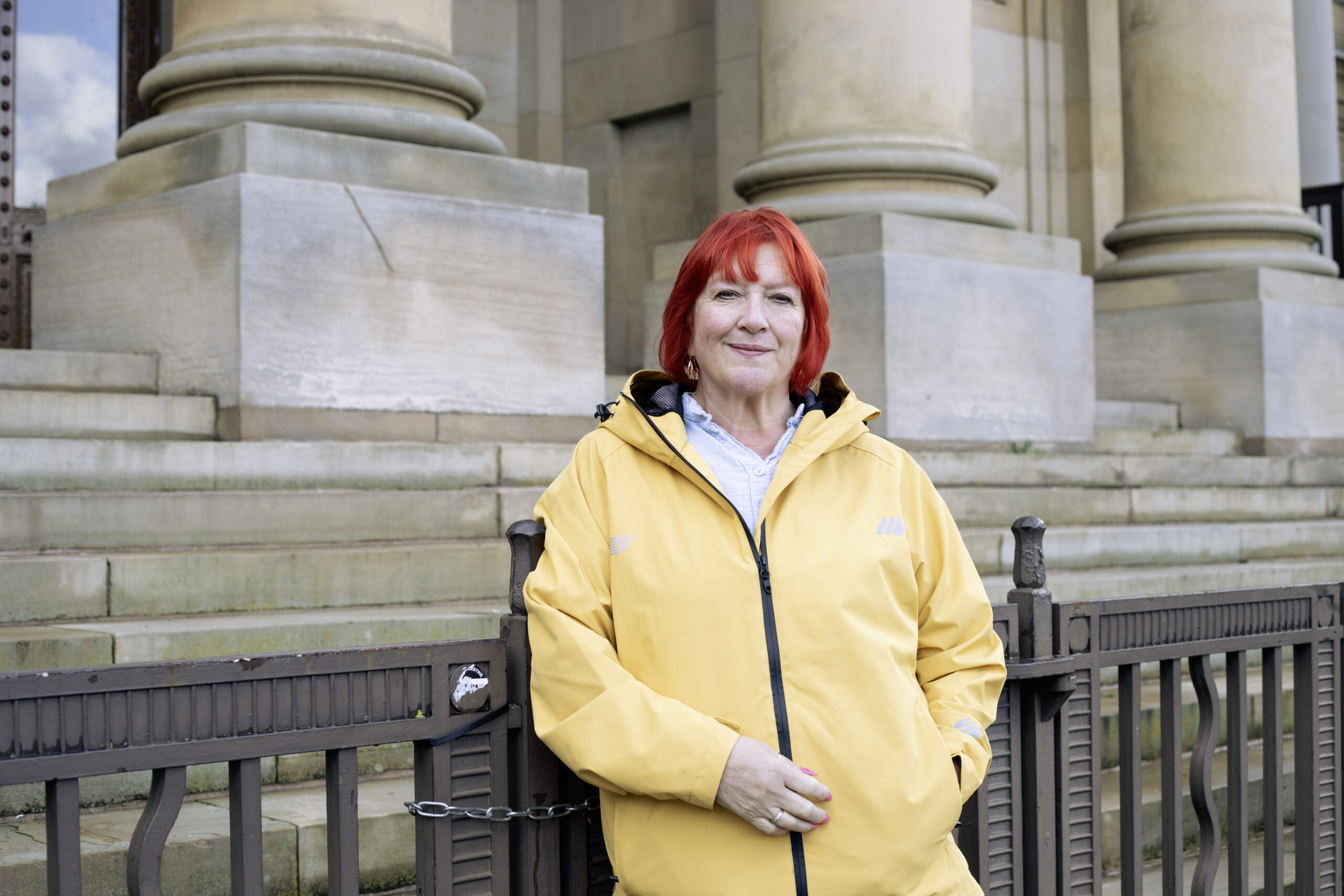 A woman with red hair and a yellow raincoat stands in front of railings in front of a neoclassical building