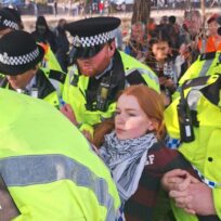 A woman with red hair, wearing a keffiyeh, is dragged off by police