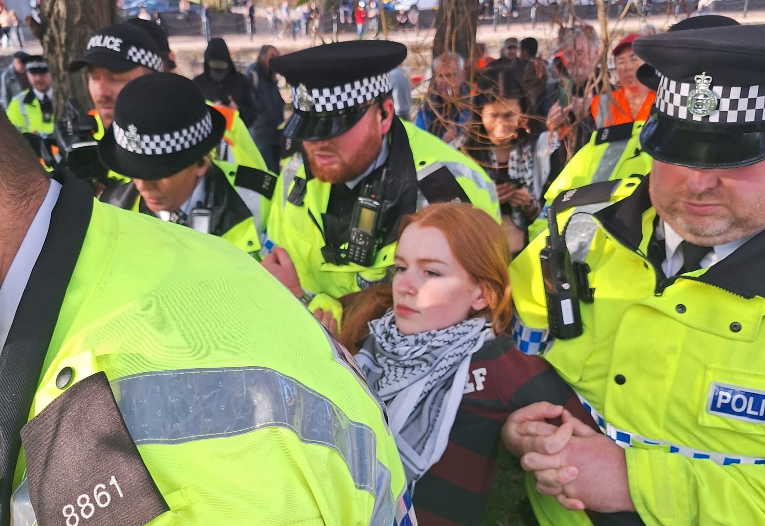 A woman with red hair, wearing a keffiyeh, is dragged off by police