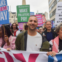 A man in a green jacket and white t-shirt holds a banner alongside other activists