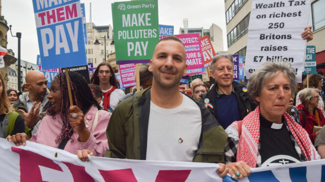 A man in a green jacket and white t-shirt holds a banner alongside other activists