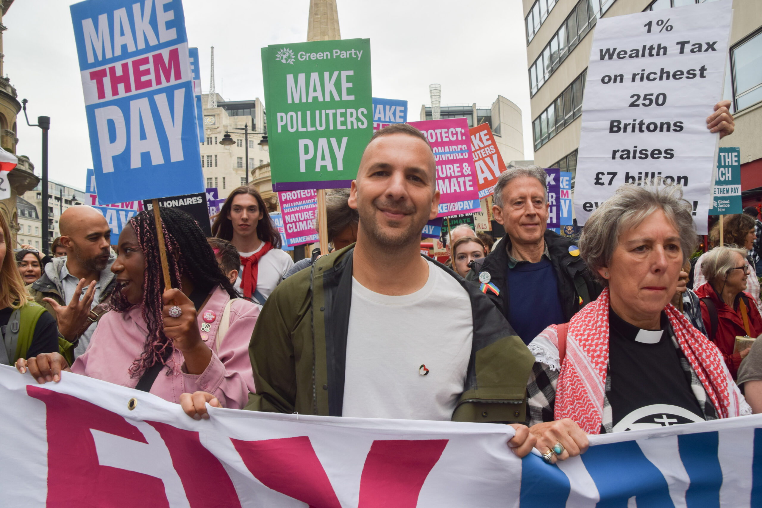 A man in a green jacket and white t-shirt holds a banner alongside other activists